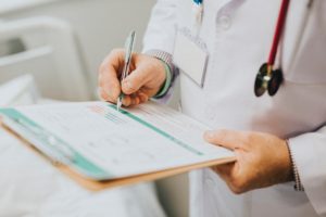 A doctor is writing on a clipboard patient chart in a hospital room.