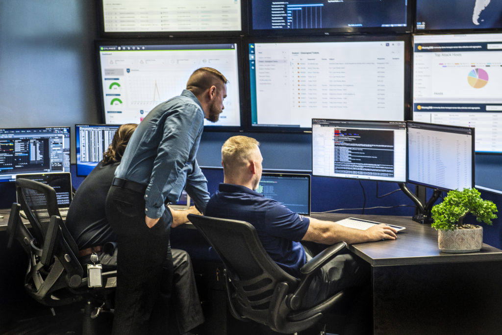 MegaplanIT Security and Compliance Services showing three staff in front of numerous computer monitors on a desk and mounted on the wall.